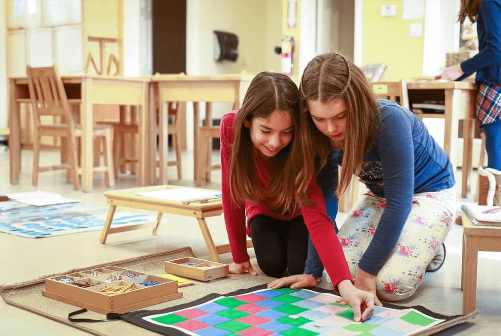 Young girls sitting on a brown mat, happily playing with a color checkered board at Mountain West Montessori in El Paso