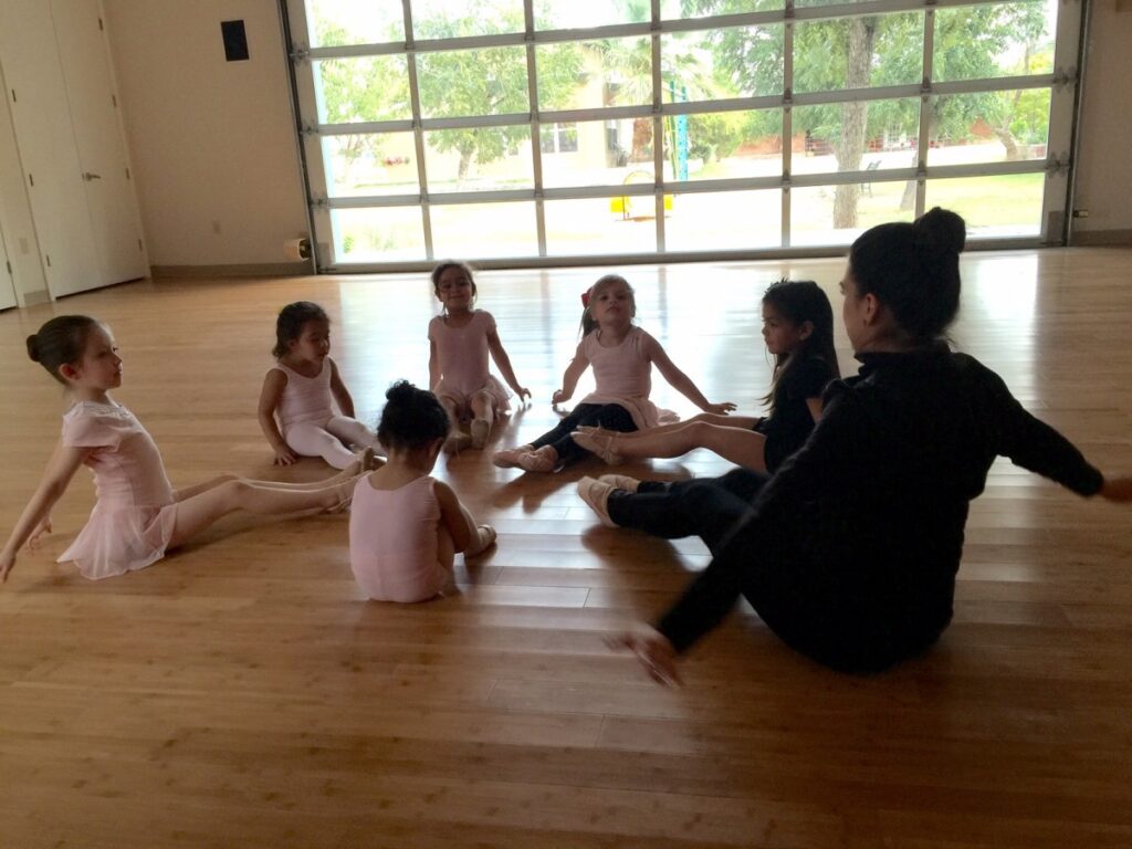 A teacher is discussing a valley dance with a group of students gathered around, all sitting on the classroom floor inside Mountain West Montessori