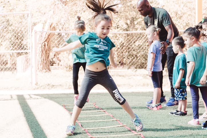 A little girl in a green Mountain West Montessori shirt is performing outdoor activities with her teacher and other classmates