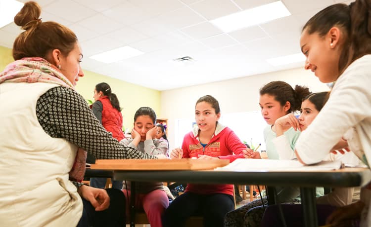 A teacher is discussing a subject with a group of students gathered around the desk.