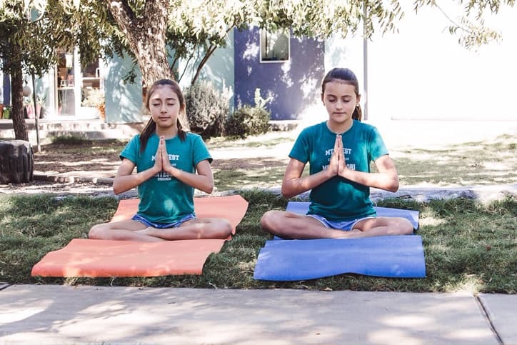 Two young girls in green Montessori T-shirts sitting on yoga mats on grass while practicing yoga