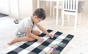 Little boy in a white dress sitting on a black and white checkered mat, playing with color blocks at Mountain West Montessori in El Paso