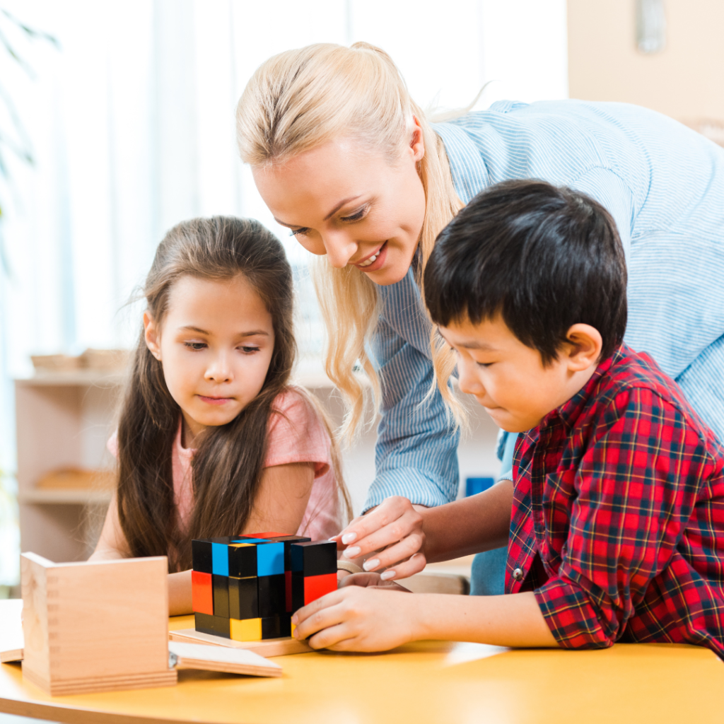 two young children, a girl and a boy, engaged in solving a large, colorful cube puzzle. A smiling adult, likely a teacher or caregiver, is leaning over them, offering guidance and encouragement. The girl, with long brown hair, watches attentively while the boy, dressed in a red plaid shirt, works on the puzzle. The setting appears to be a classroom with a calm and supportive learning environment, featuring wooden materials on the table. The teacher's posture suggests a nurturing and hands-on approach to facilitating learning.